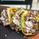 Three dirty steak tacos assembled with skirt steak, guacamole, chipotle sauce and corn tortillas and lined up on a cutting board