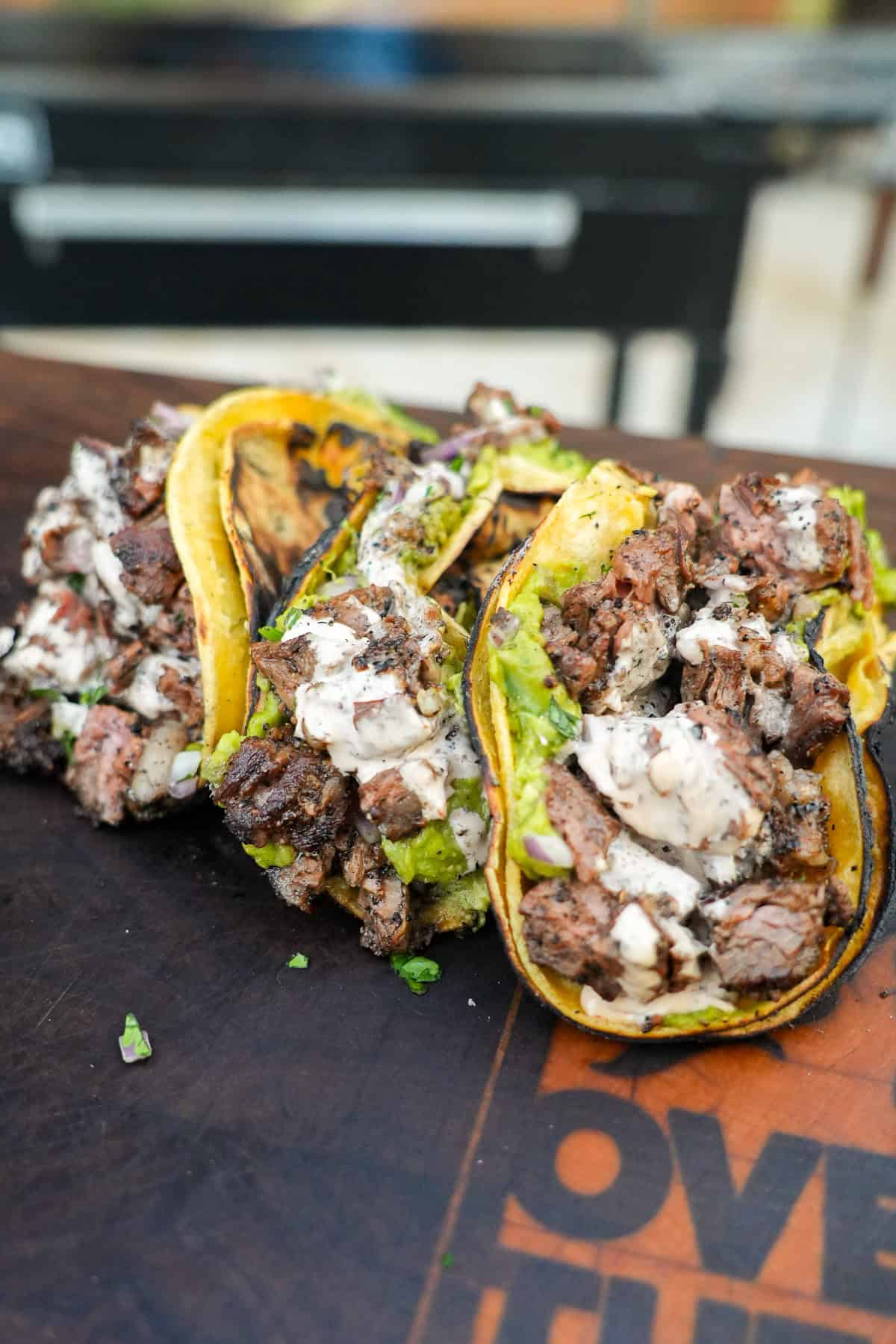 Three dirty steak tacos assembled with skirt steak, guacamole, chipotle sauce and corn tortillas and lined up on a cutting board