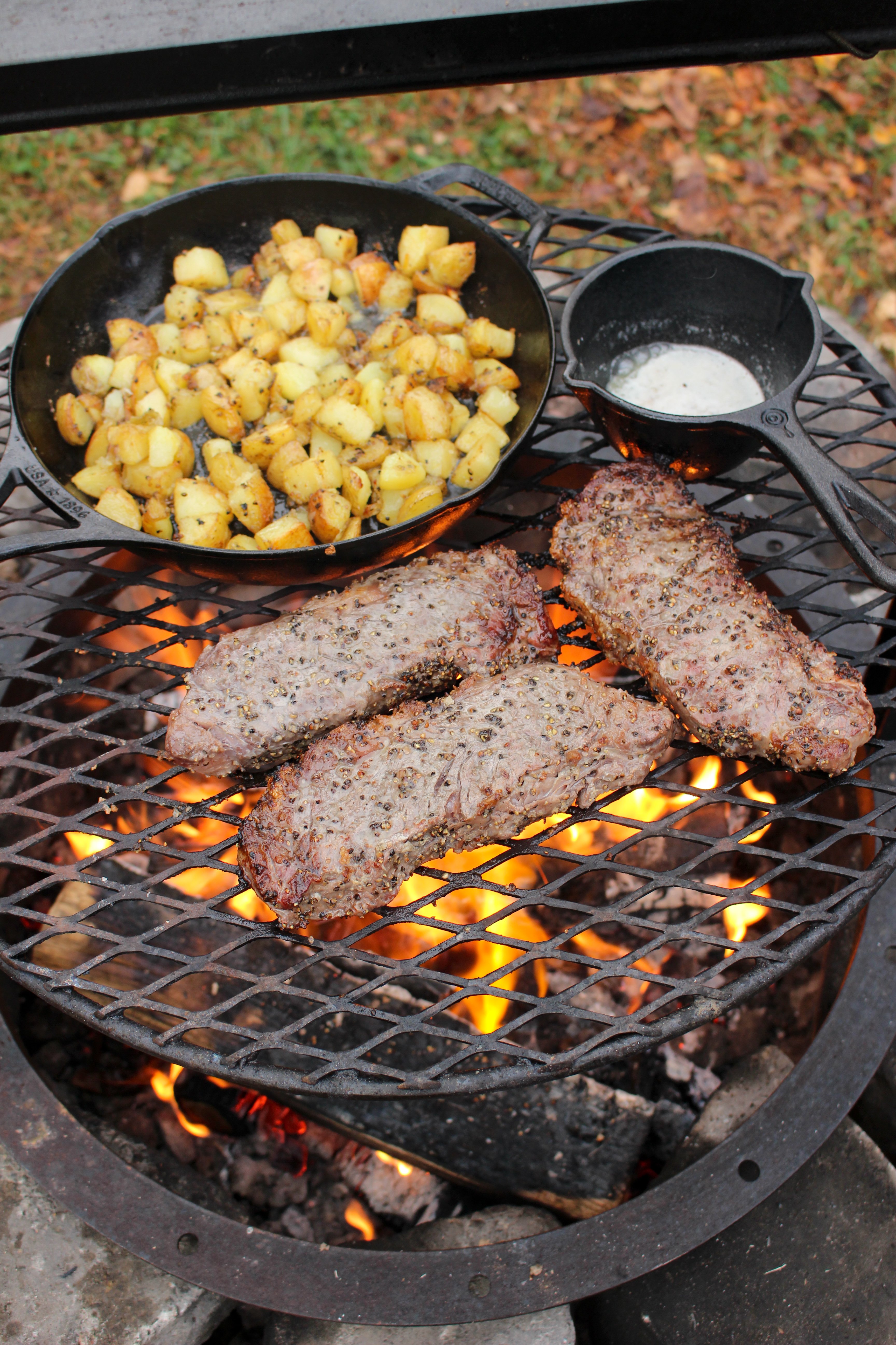 The steak, potatoes and butter all sitting on the grill grate over the flames together.