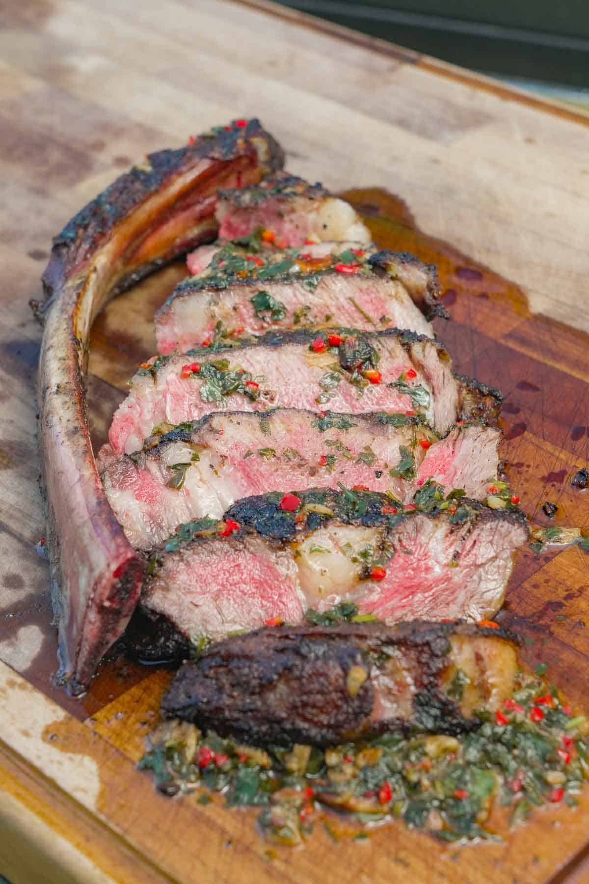 Sliced tomahawk steak drizzled with chimichurri sauce on a cutting board