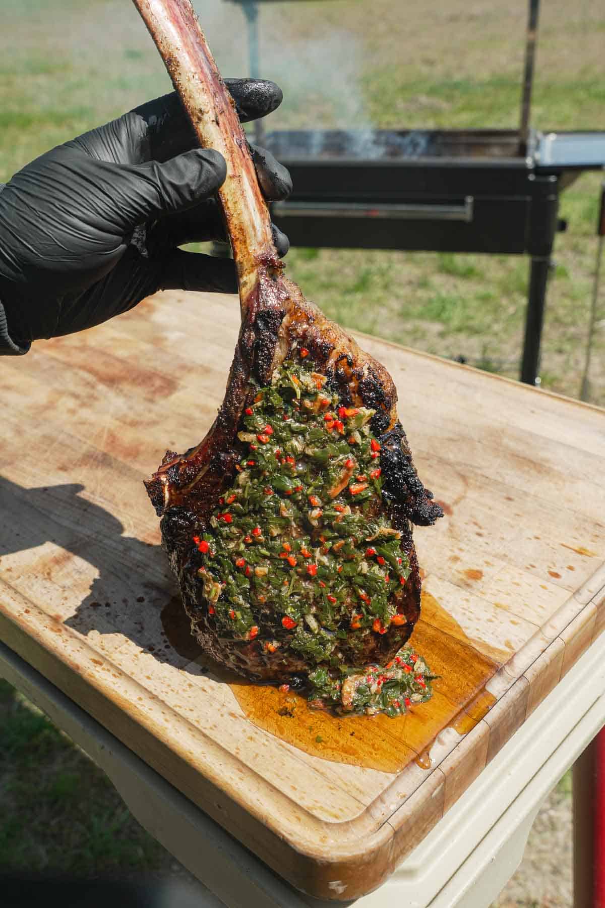 A hand holding the hanging tomahawk steak with garlic confit chimichurri by the long bone on a cutting board