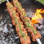 Spiced Lamb Kebabs with Mint Chimichurri on the grill with flames in the background.