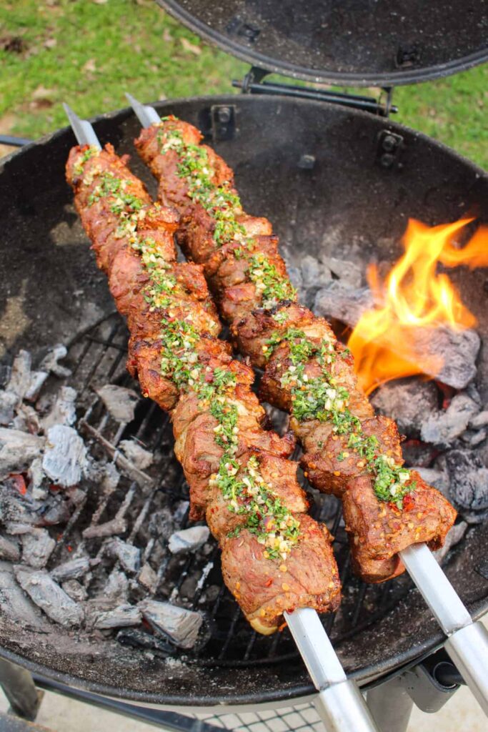 Spiced Lamb Kebabs with Mint Chimichurri on the grill with flames in the background.