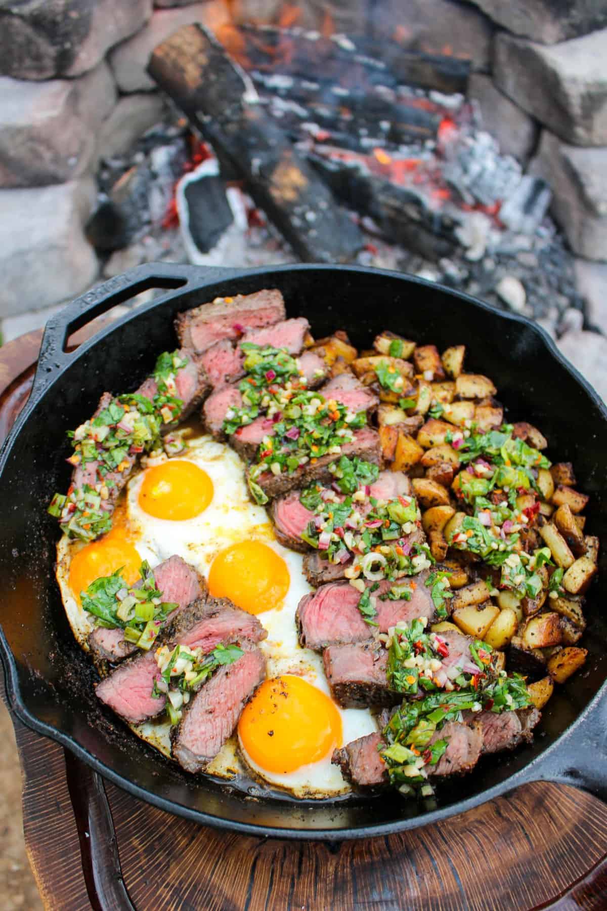 overhead of a skillet filled with steak and eggs with charred scallion chimichurri next to a fire pit