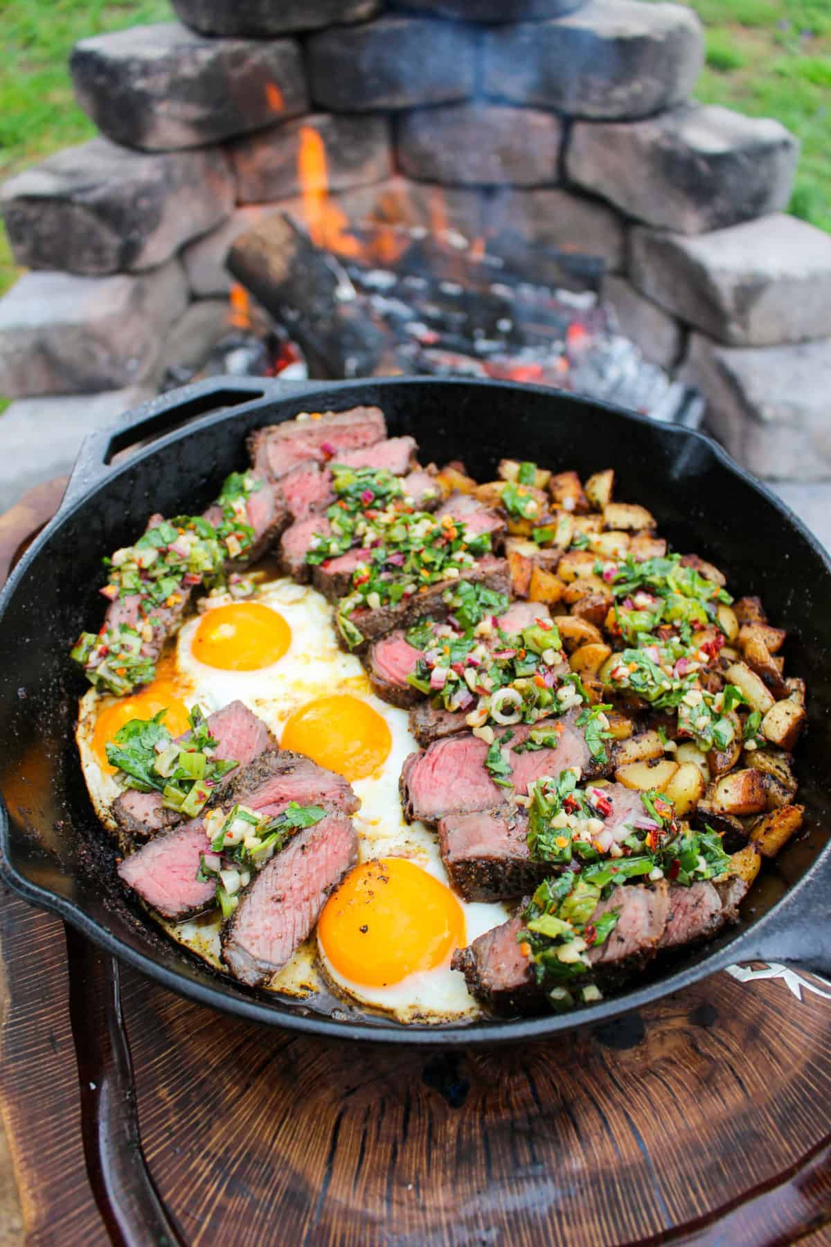 a skillet next to a fire filled with fried eggs, sliced steak, and potatoes topped with charred scallion chimichurri