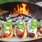 five bacon wrapped chorizo dogs topped with cream sauce and jalapeño in front of a fire pit