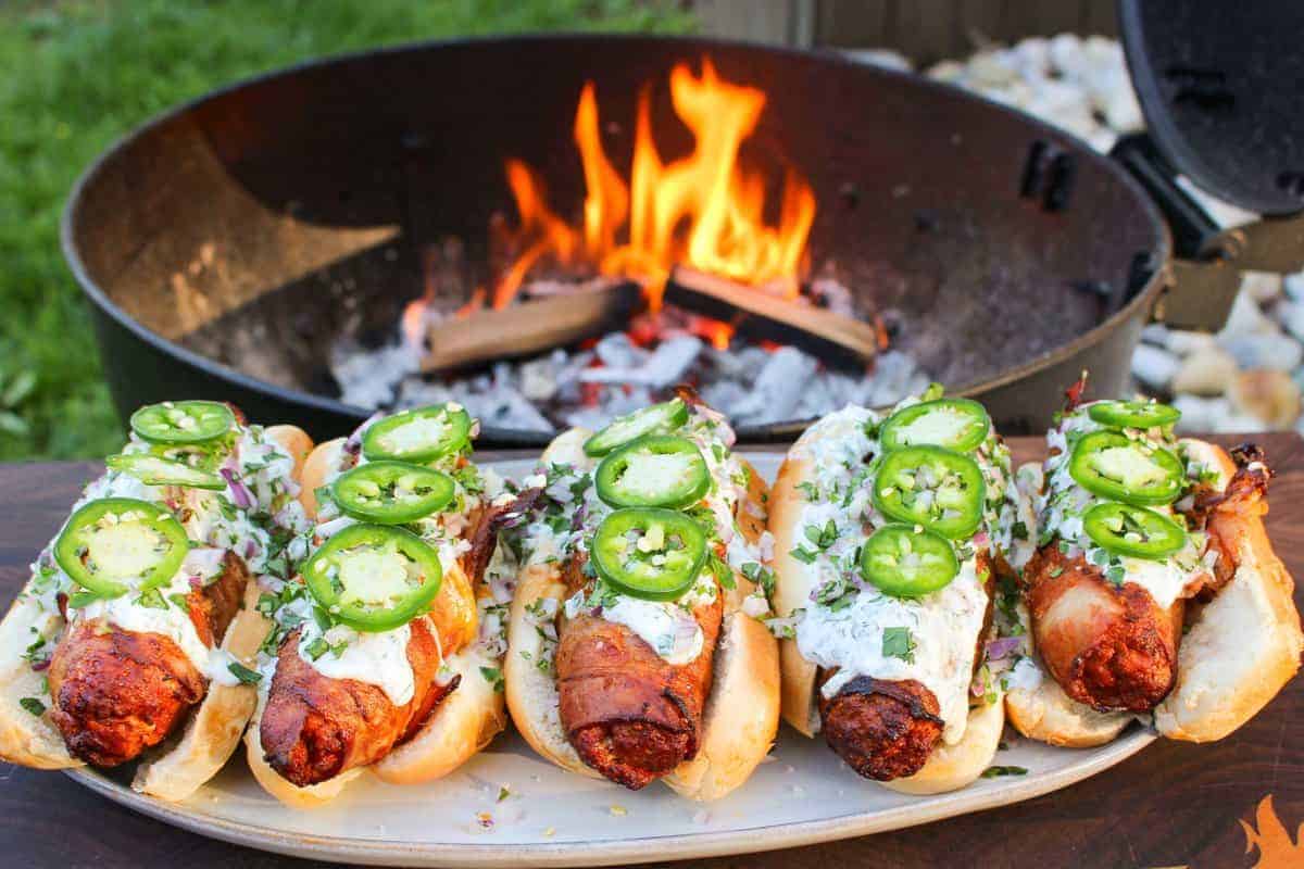 five bacon wrapped chorizo dogs topped with cream sauce and jalapeño in front of a fire pit