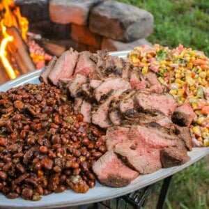 large plate filled with sliced tri-tip, pinquito beans, and charred corn salsa held over a fire