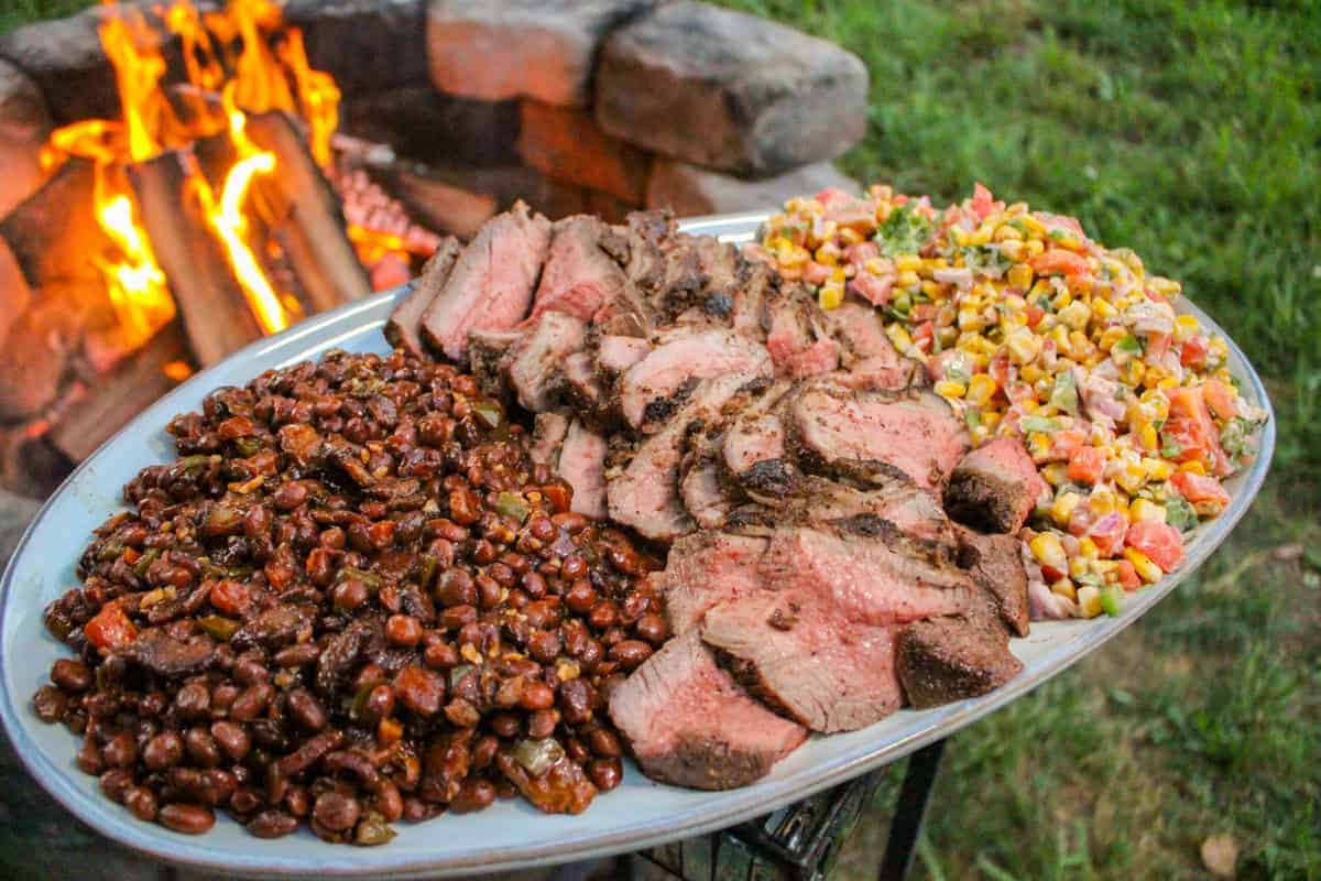 large plate filled with sliced tri-tip, pinquito beans, and charred corn salsa held over a fire