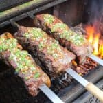 three strip steak skewers topped with salsa vinaigrette over a fire