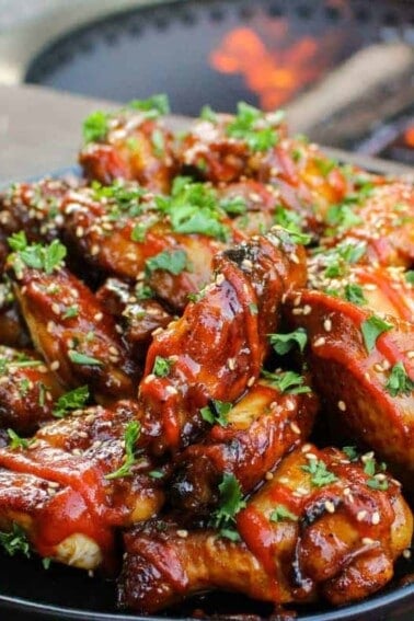 a large plate piled with huli huli chicken wings and garnished with sesame seeds, sriracha, and cilantro