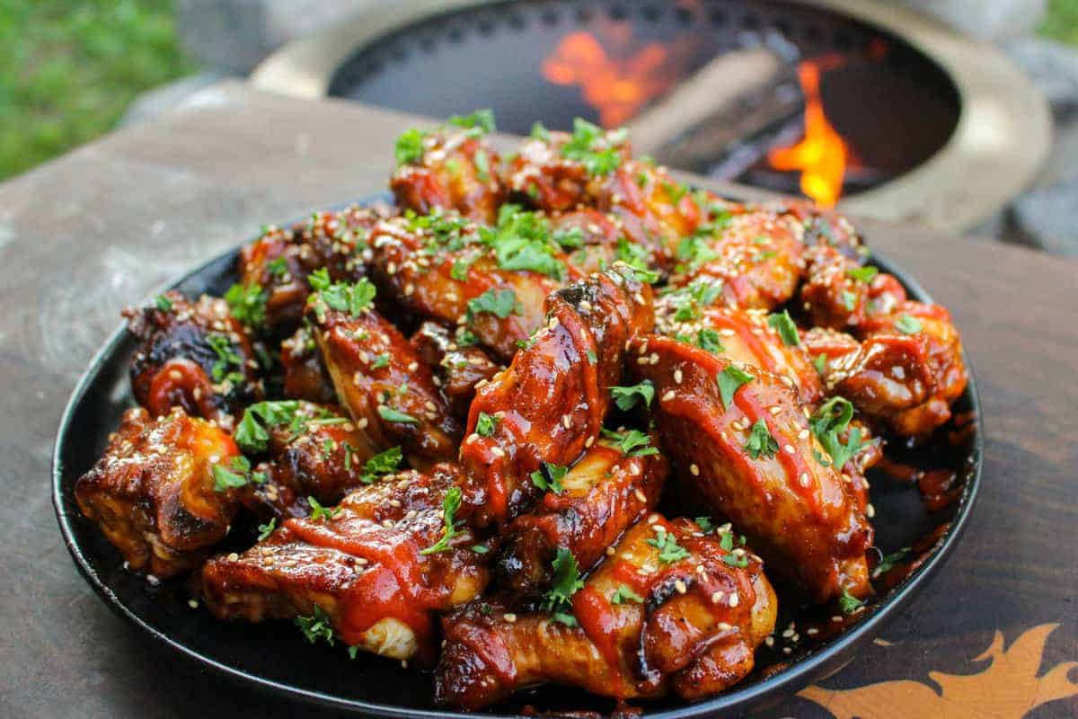 a large plate piled with huli huli chicken wings and garnished with sesame seeds, sriracha, and cilantro