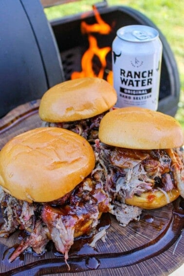 side view of three pulled pork sandwiches next to a smoker and a can of Ranch Water