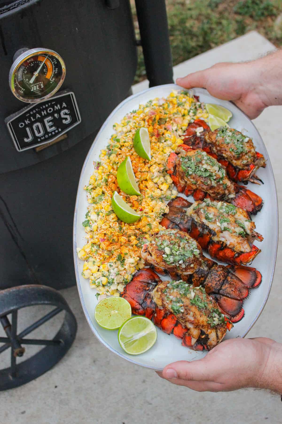 a platter with smoked lobster tails and esquites held next to a smoker