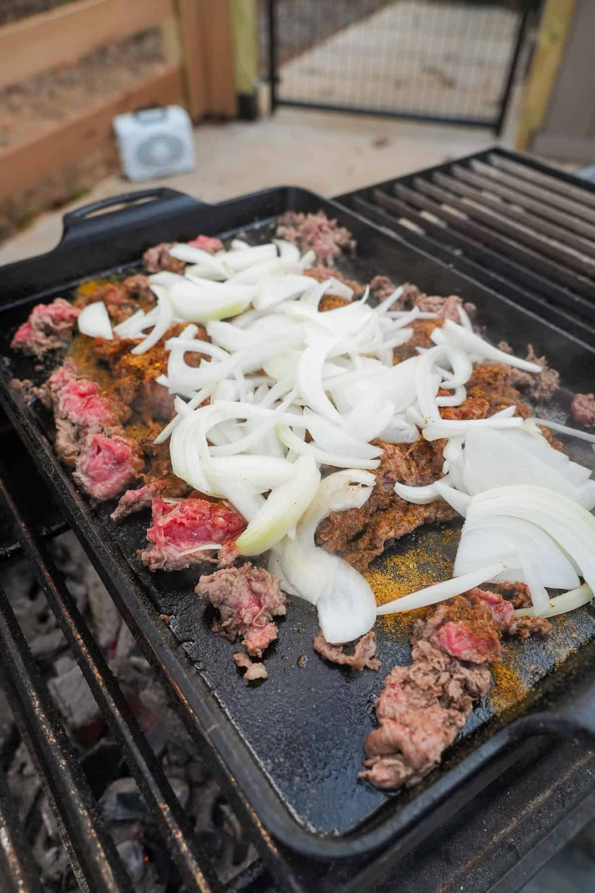 Grilling thinly sliced ribeye and sliced white onions on a cast iron plancha for pizza cheesesteak