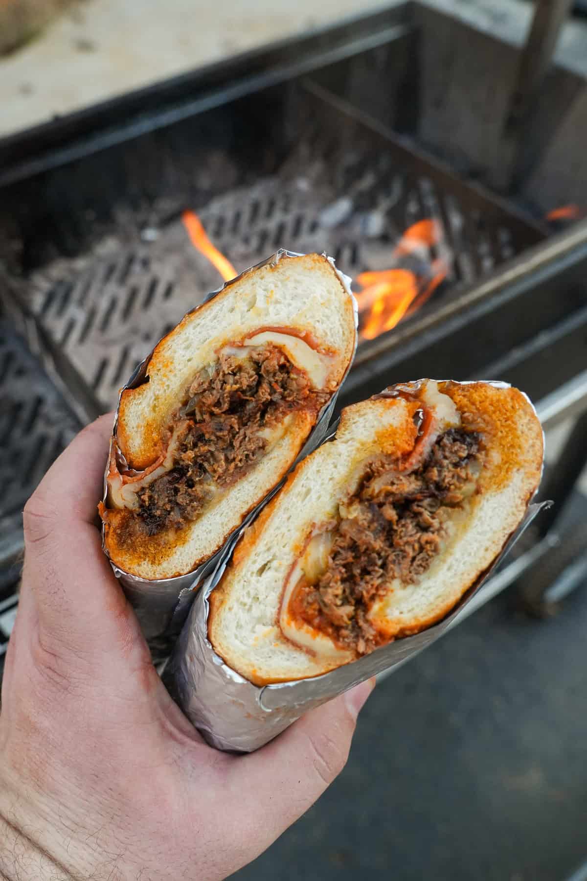 Derek Wolf holding a sliced pizza cheesesteak sandwich to show inner ingredients, in front of a FYR Grill