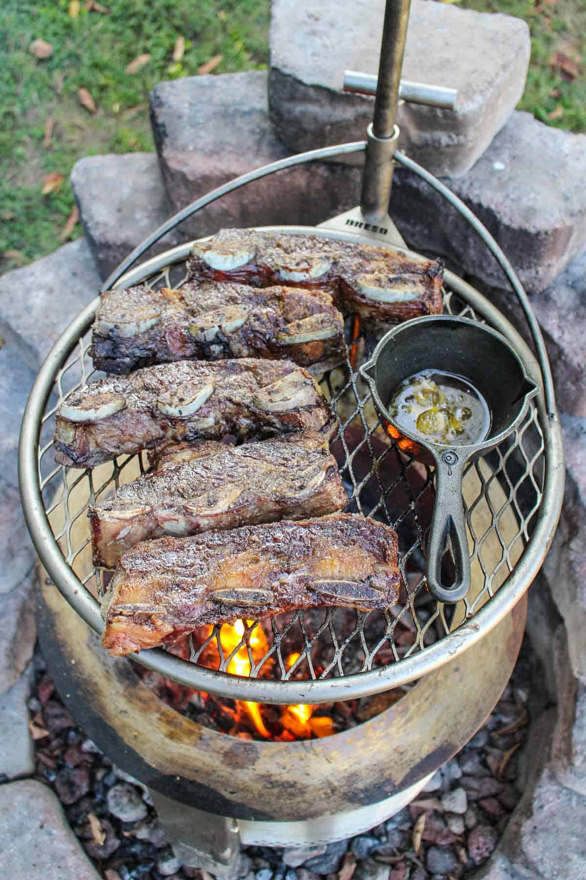 Grilled steak on a grill next to a skillet of basting liquid.