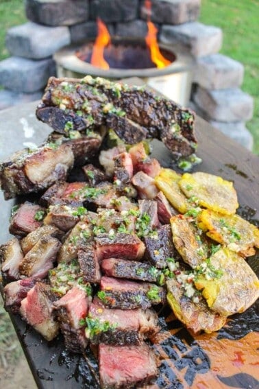 closeup of sliced steak topped with chimichurri butter next to smashed potatoes on a cutting board