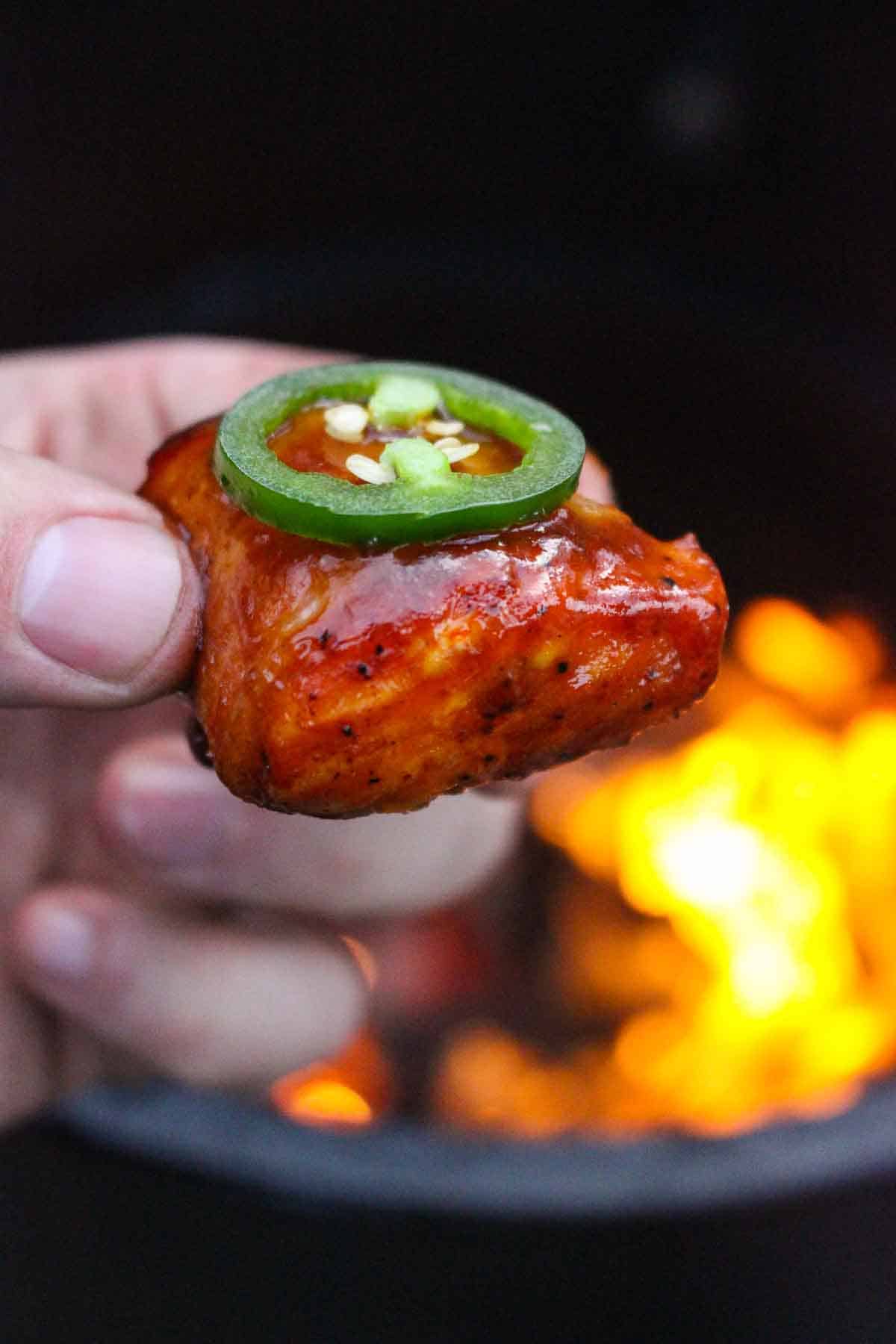 One of the Maple Bacon Bourbon Salmon Bites being held close to the camera with the fire in the background.
