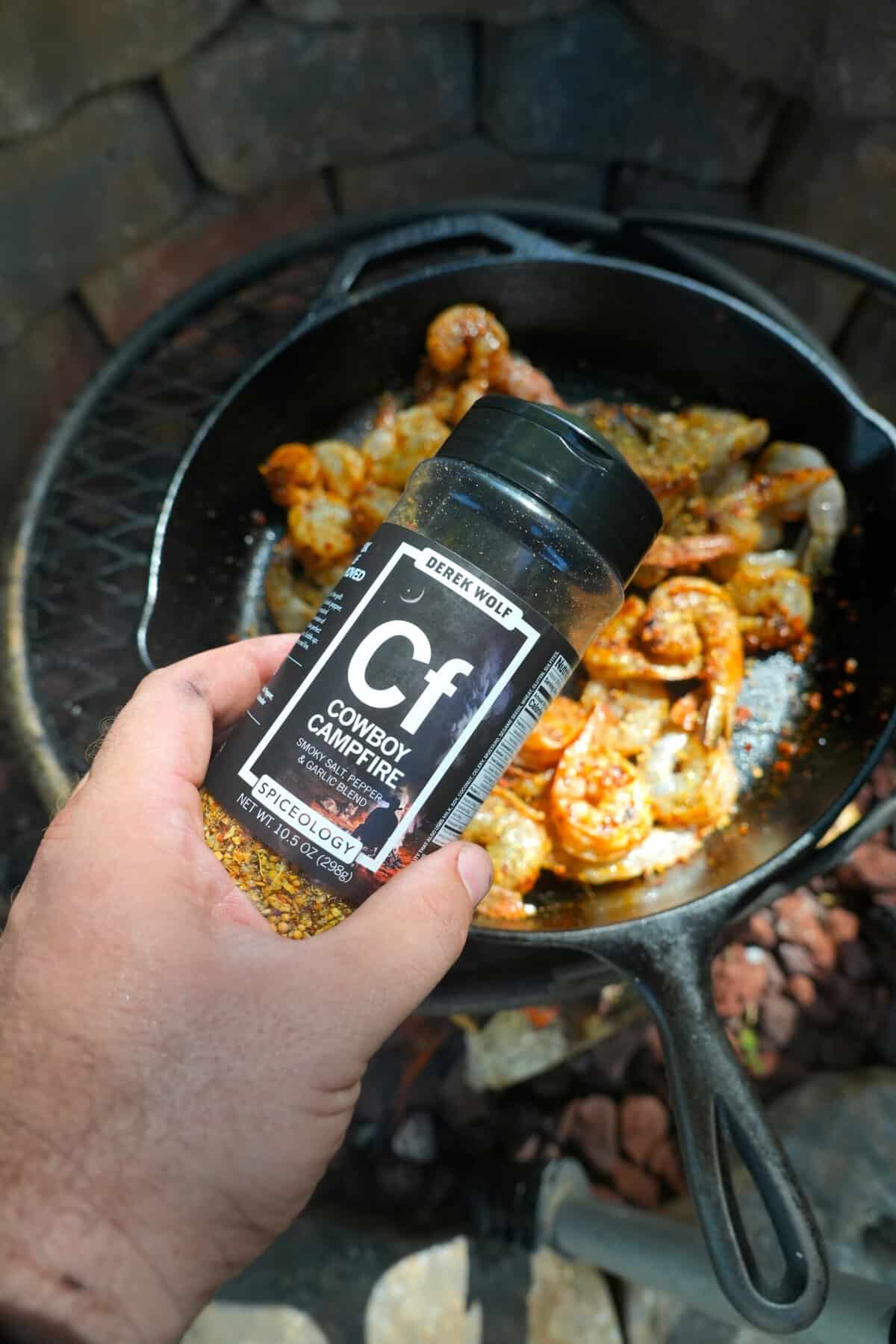 Derek Wolf holding a jar of Cowboy Campfire Seasoning, ready to add the rub to Spanish garlic shrimp in a cast iron skillet