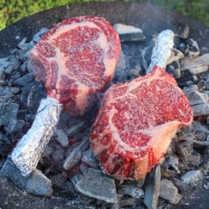Two raw tomahawk steaks cooking on the coals.