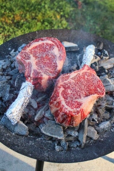 Two raw tomahawk steaks cooking on the coals.