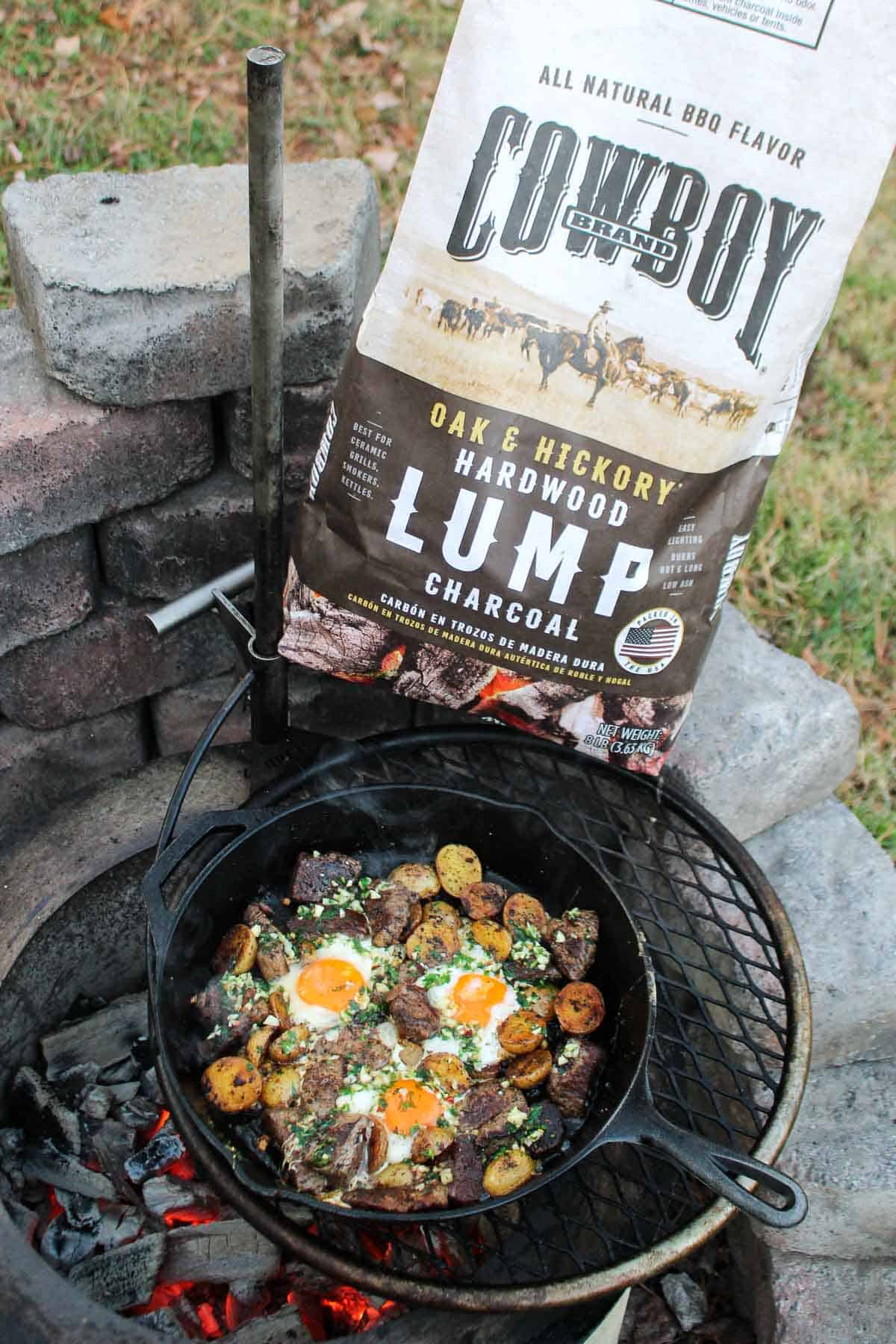 A bag of Cowboy Charcoal sits next to the fire pit with the finished steak, eggs, and potatoes. 