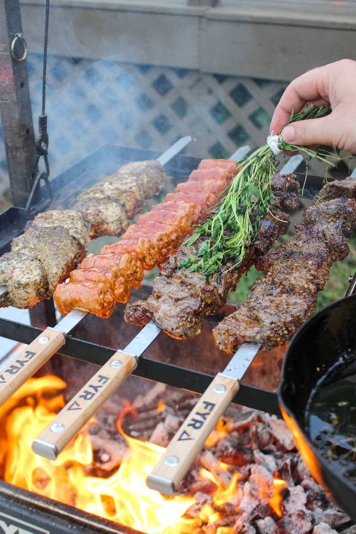 Pork, sausage and beef skewers cooking over some flames.