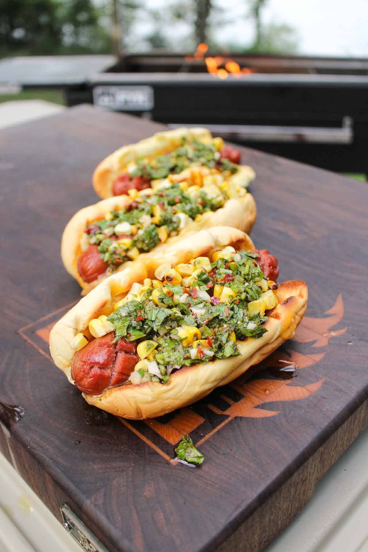 Grilled hot dogs on a cutting board, served with charred corn and chimichurri.