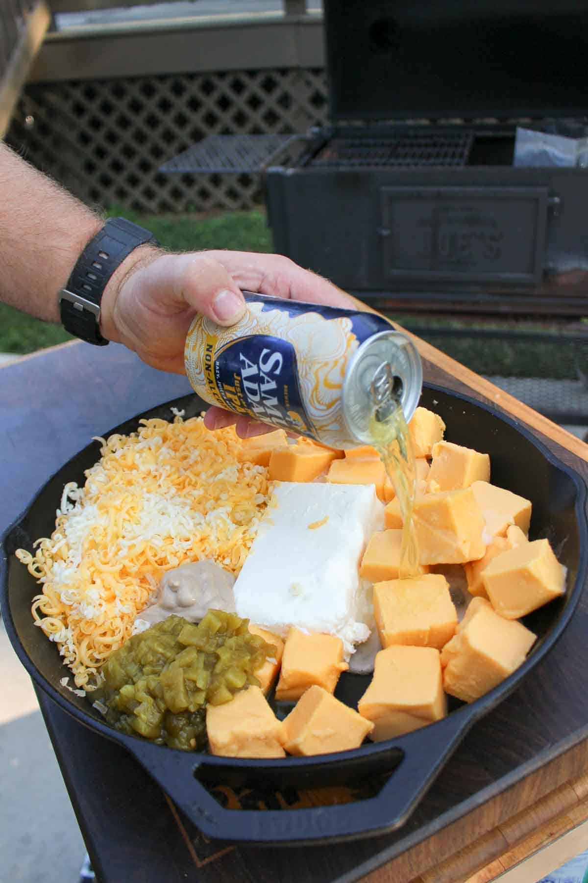 Pouring Sam Adams Just the Haze IPA into the cast iron along with the other smoked beer cheese ingredients.