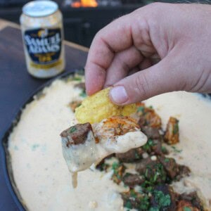 Holding a chip up to the camera that's dripping in smoked beer cheese with a bite of steak and shrimp also on the chip.