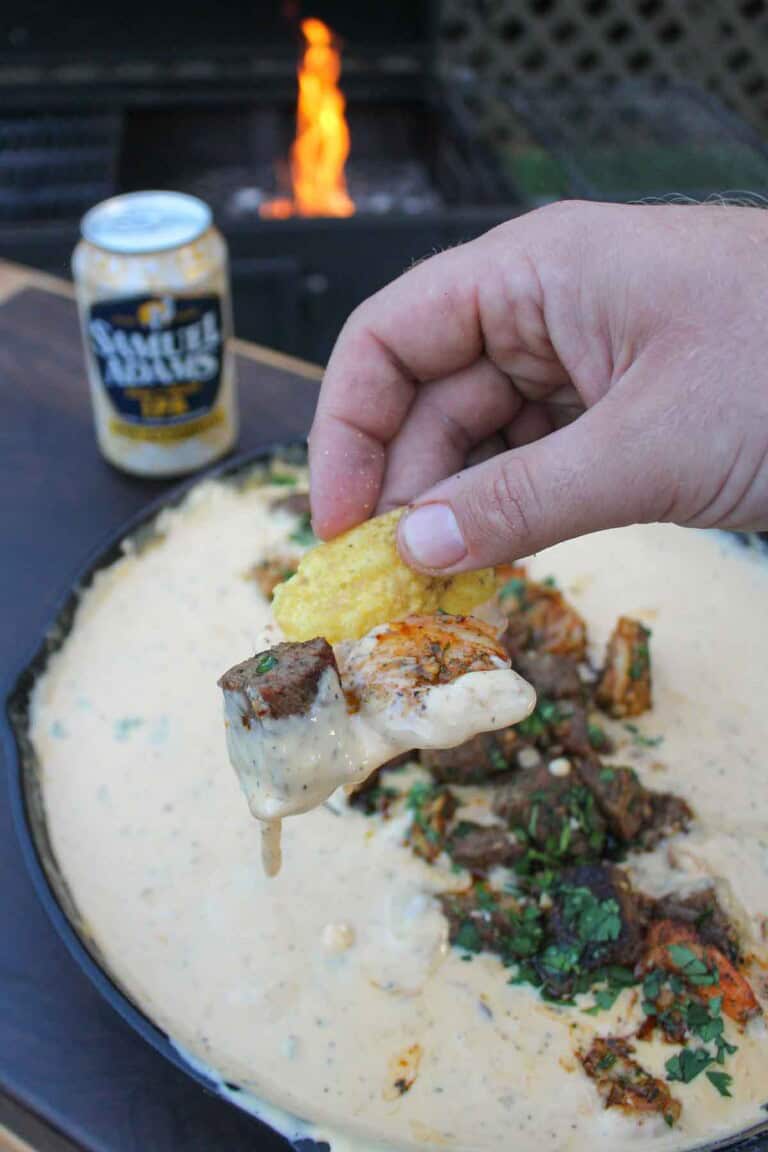 Holding a chip up to the camera that's dripping in smoked beer cheese with a bite of steak and shrimp also on the chip.