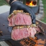 A gloved hand holding a crusted tomahawk steak sliced open to reveal the delicious crust and the juicy interior of the meat