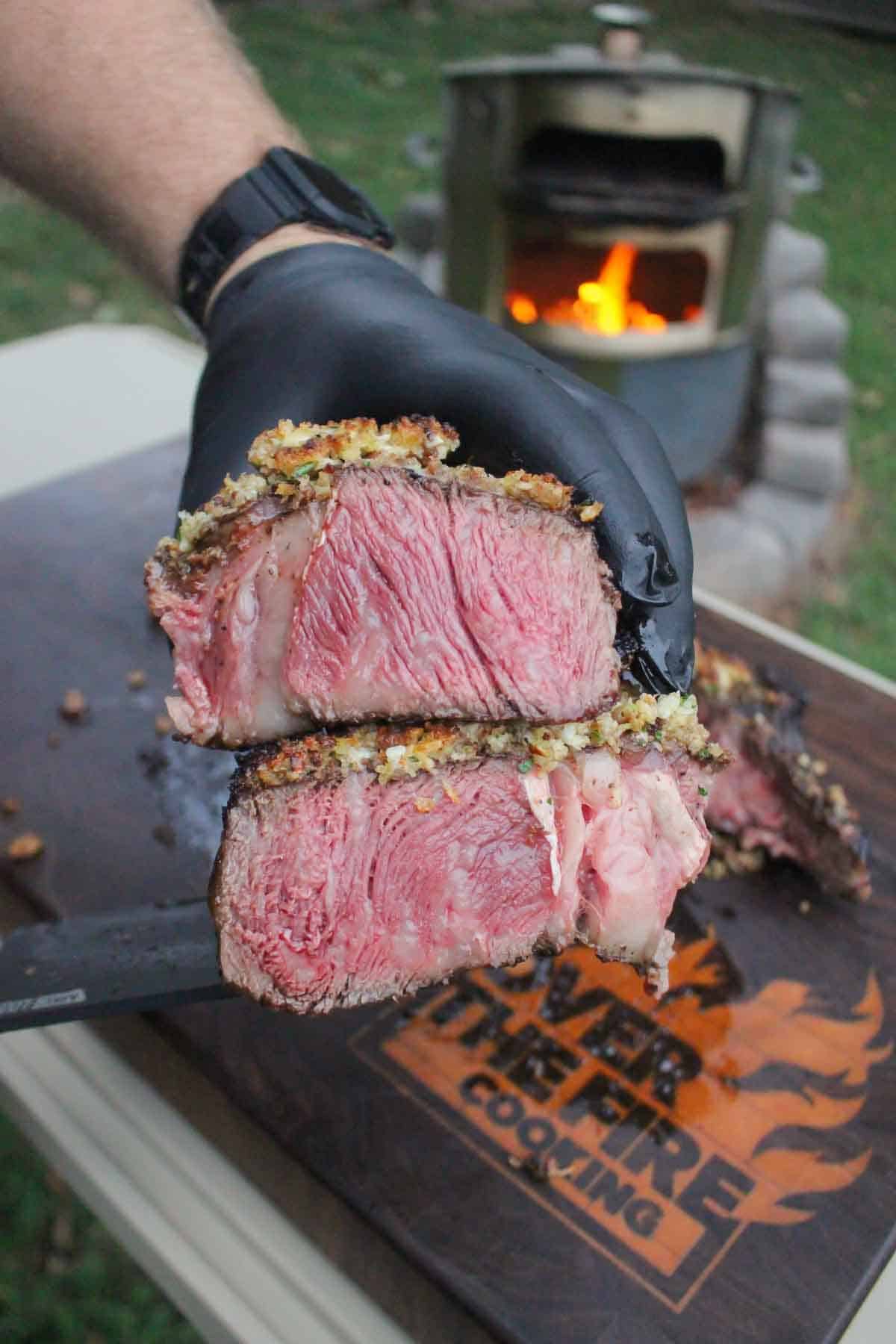 A gloved hand holding a crusted tomahawk steak sliced open to reveal the delicious crust and the juicy interior of the meat