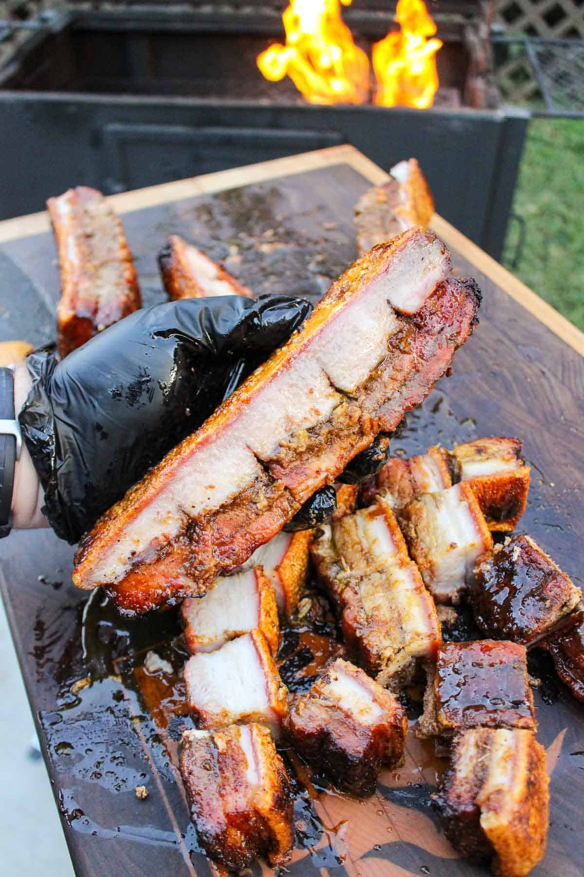 A gloved hand holding a slab of crispy pork belly burnt ends with cubes of pork belly burnt ends on a cutting board in front of a grill fire