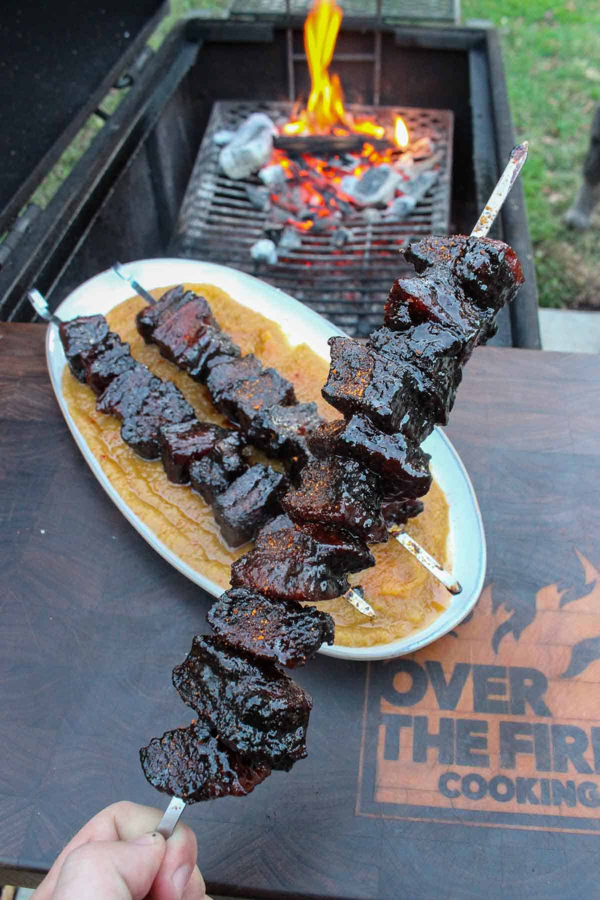 Holding up a skewer of smoked pork belly burnt ends with applesauce next to the grill