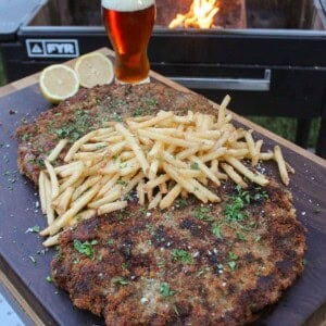 Milanesa with fries, served on a platter with a sliced lemon and a cold beer, and in front of a grill fire