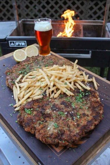 Milanesa with fries, served on a platter with a sliced lemon and a cold beer, and in front of a grill fire