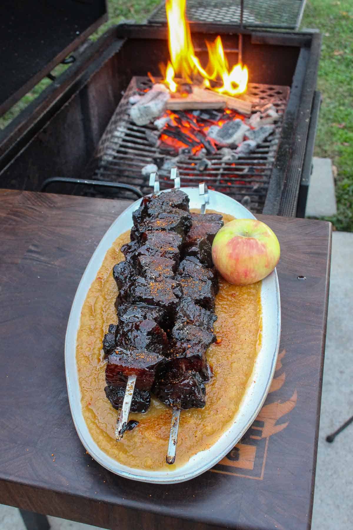 Smoked pork belly burnt ends with applesauce on a platter next to the grill, with an apple next to the dish