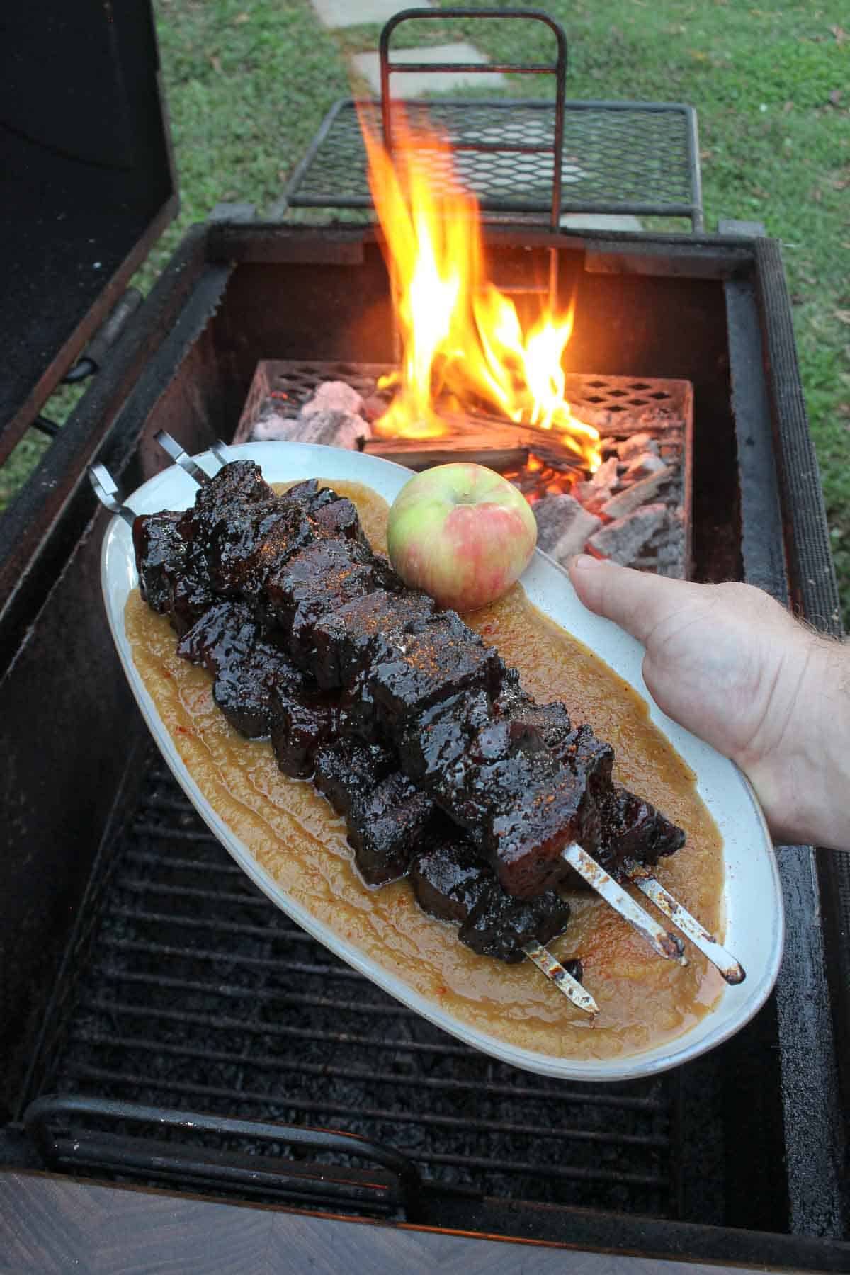 Smoked pork belly burnt ends with applesauce, held next to a grill fire with apples on the side