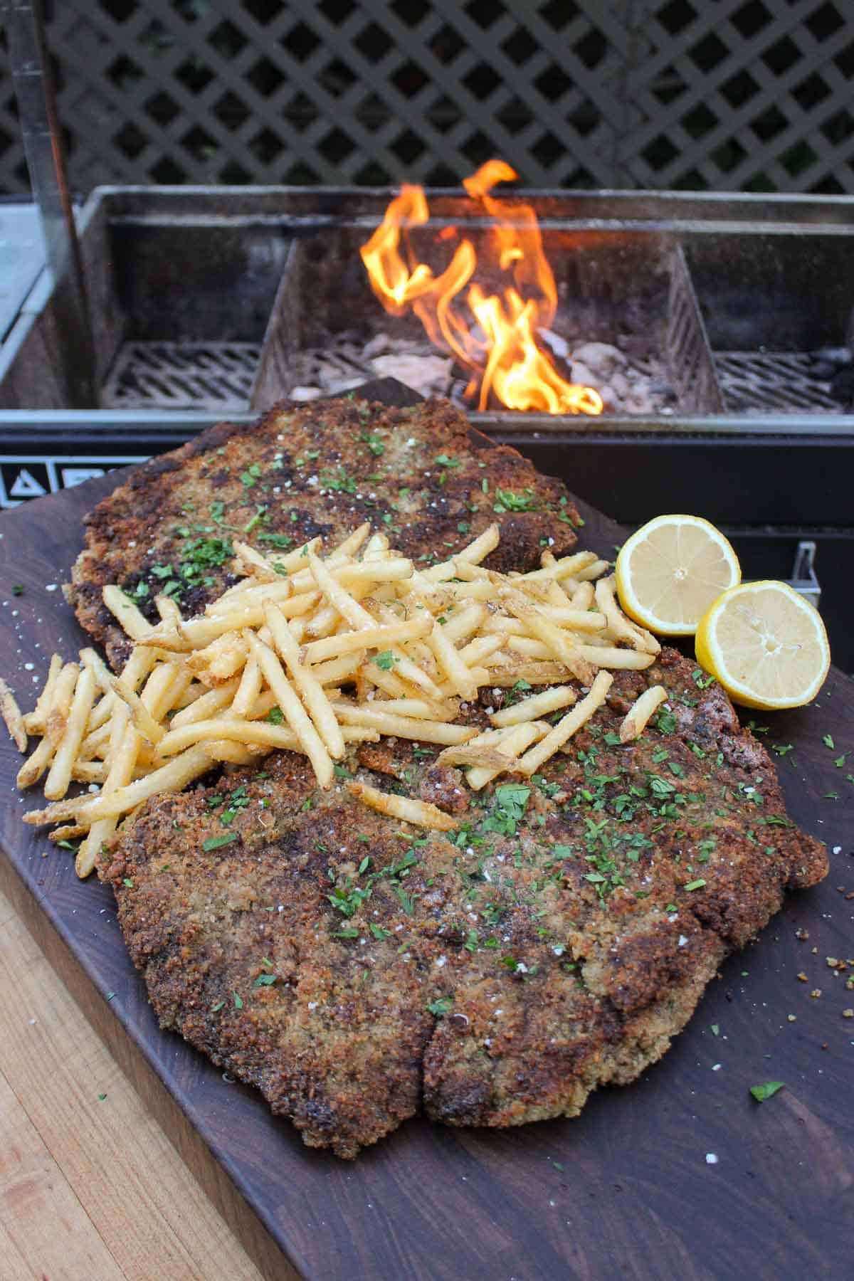 Milanesa with fries on top, served with a sliced lemon and in front of a roaring grill fire