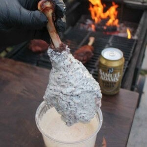 A gloved hand dipping a beer brined turkey leg in Alabama white sauce with Samuel Adams Winter Lager and a grill fire in the background