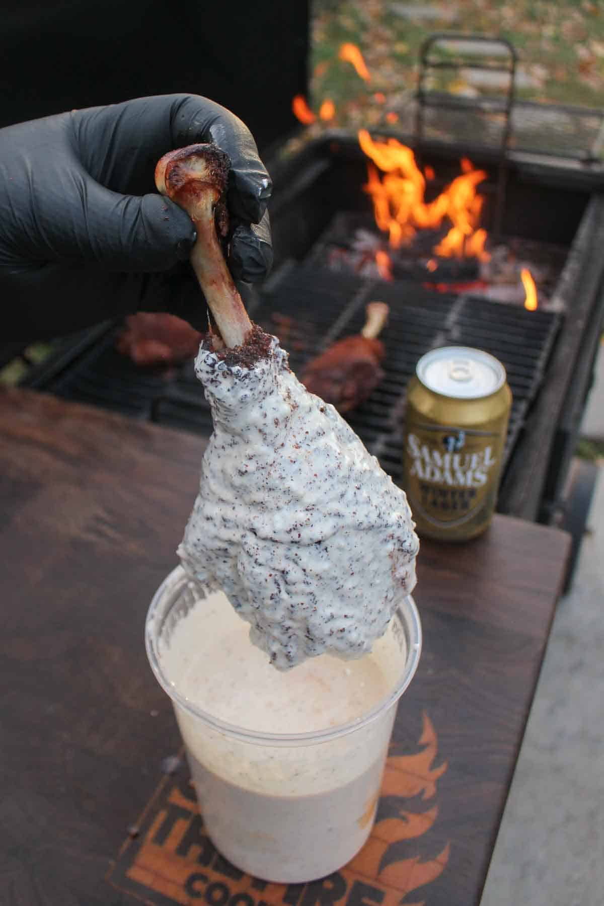 A gloved hand dipping a beer brined turkey leg in Alabama white sauce with Samuel Adams Winter Lager and a grill fire in the background