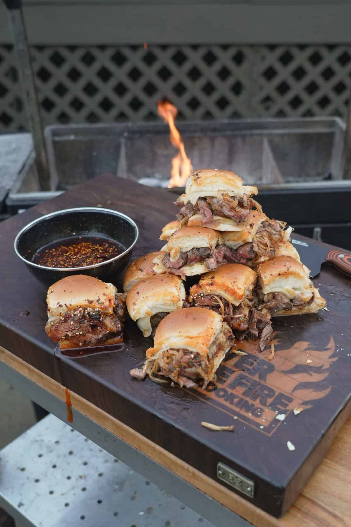 Hot honey steak sliders piled high on a cutting board with hot honey butter for dipping on the side and a grill in the background