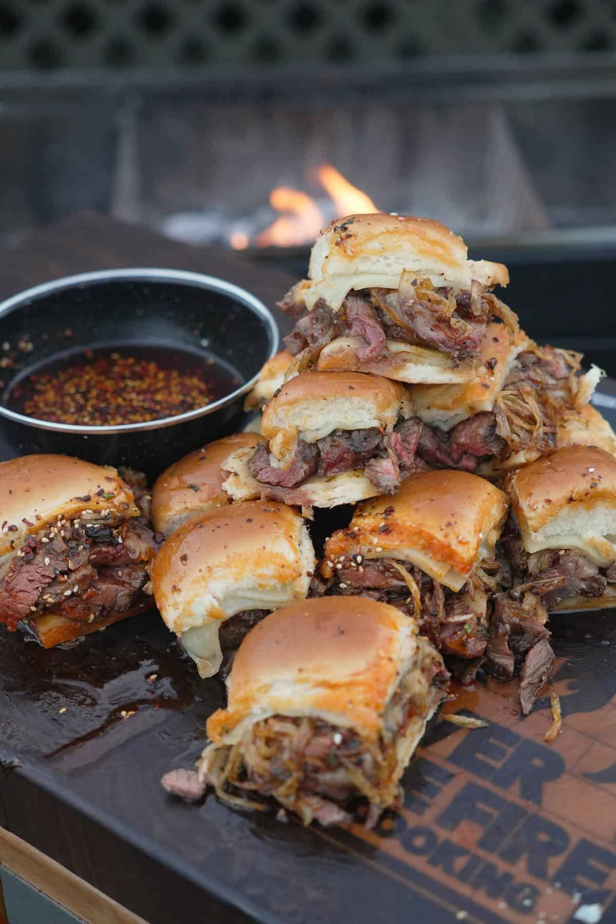 A pile of hot honey steak sliders with crispy onions next to the grill, with more hot honey for dipping on the side