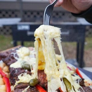 A hand holding a fork pulling raclette cheese from a platter of grilled raclette: steak, sausages, cheese, potatoes, baguette, pickles, pearl onions and red peppers