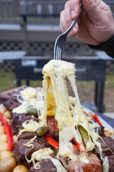 A hand holding a fork pulling raclette cheese from a platter of grilled raclette: steak, sausages, cheese, potatoes, baguette, pickles, pearl onions and red peppers