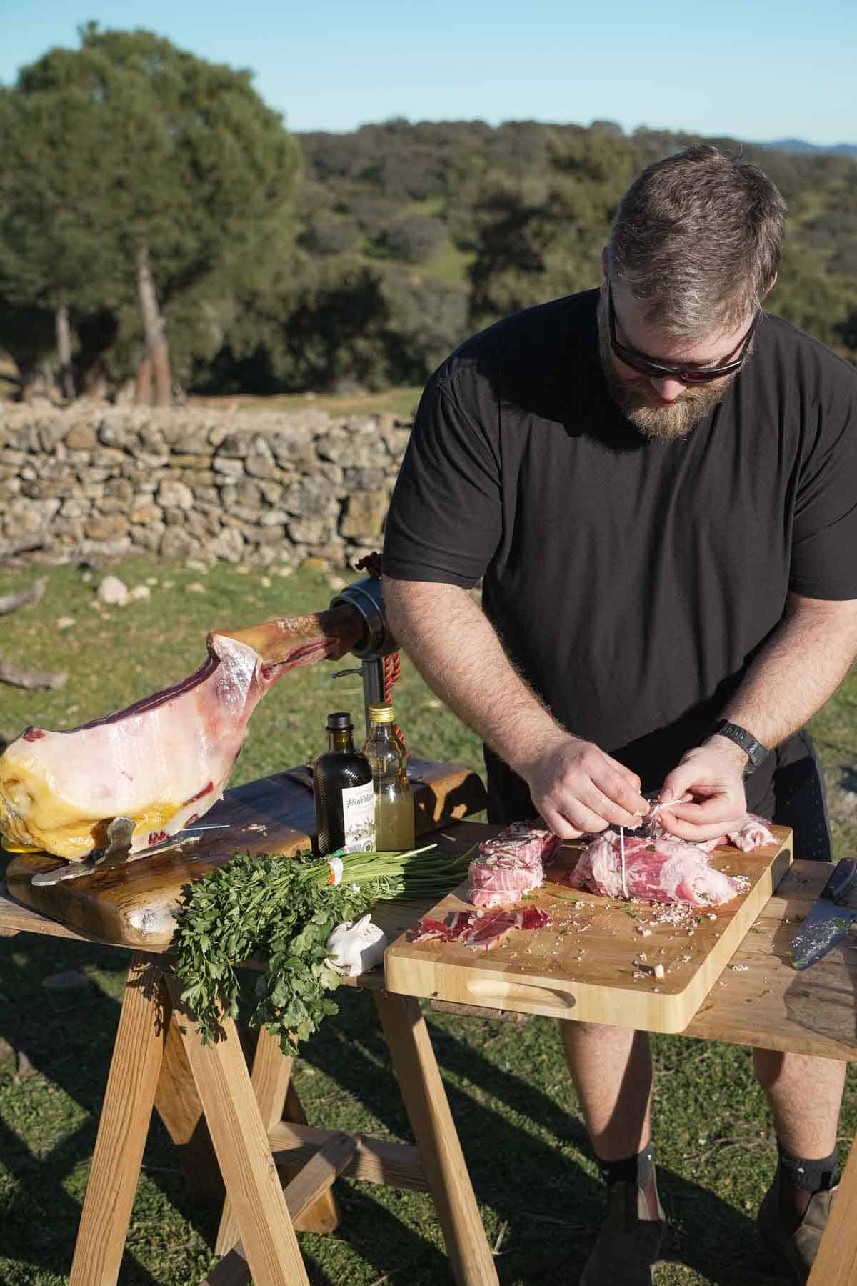 Derek Wolf assembling pork pinwheels on a wooden cutting board outside with a pork leg next to him