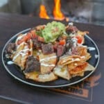 A plate of fried nachos with quesadilla chips, skirt steak, guacamole and other nacho toppings in front of a grill fire
