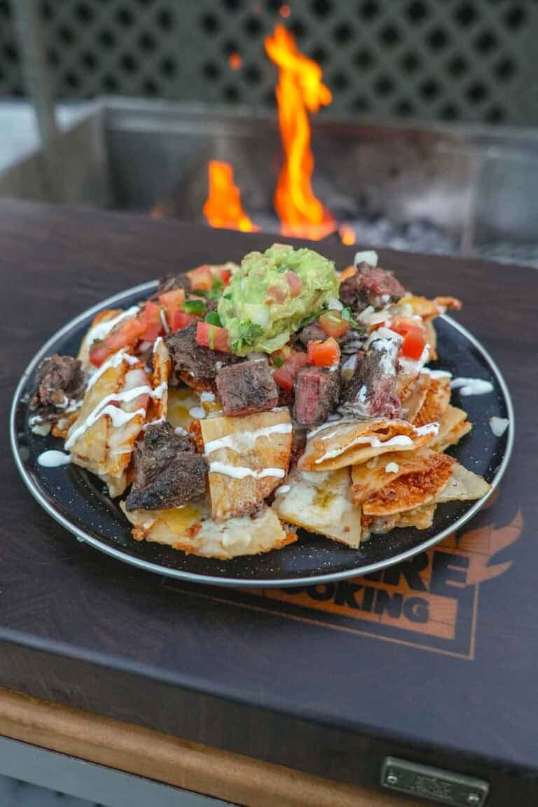 A plate of fried nachos with quesadilla chips, skirt steak, guacamole and other nacho toppings in front of a grill fire